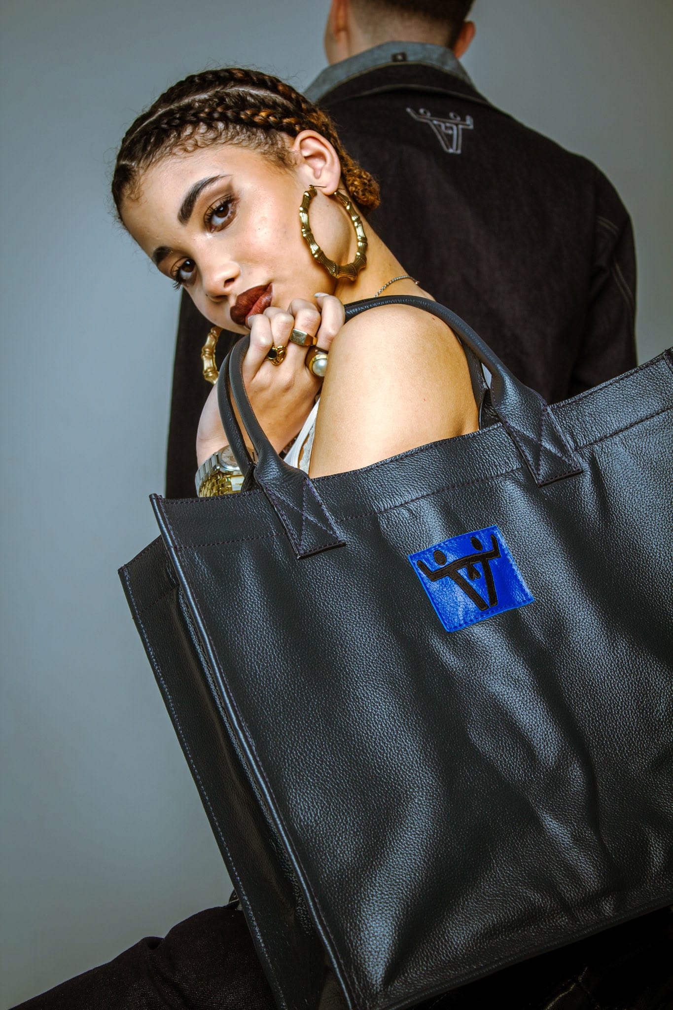 Femme aux tresses et créoles dorées portant un grand sac noir au logo Chrysalis bleu