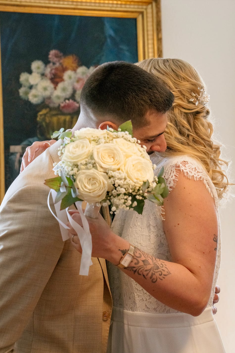 Kenza et Steven enlacés derrière un bouquet de roses blanches, devant un tableau de fleurs