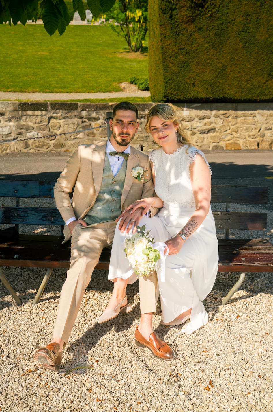 Kenza et Steven assis sur un banc au soleil, le bouquet sur les genoux, devant un mur de pierre