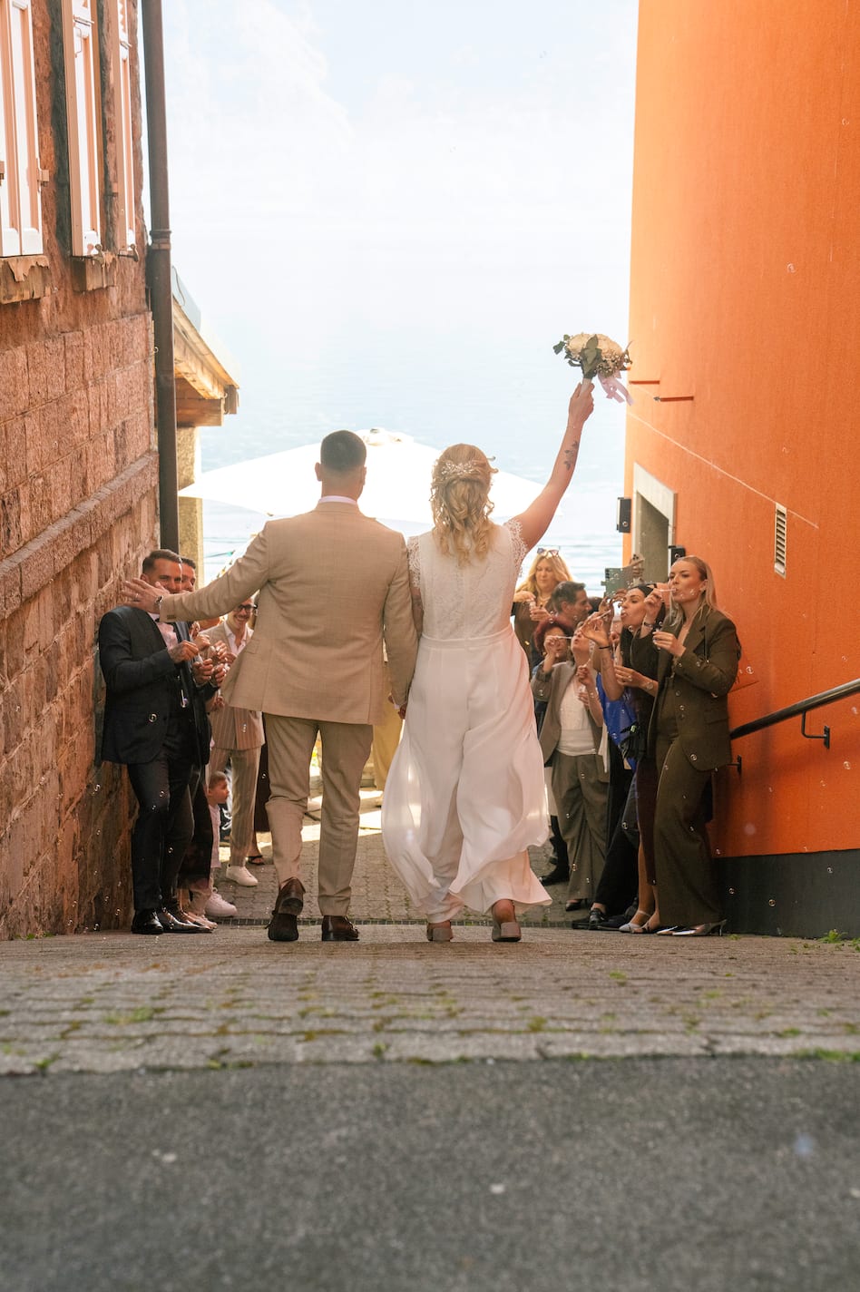 Kenza et Steven, de dos, remontent une ruelle entre leurs invités, bouquet levé