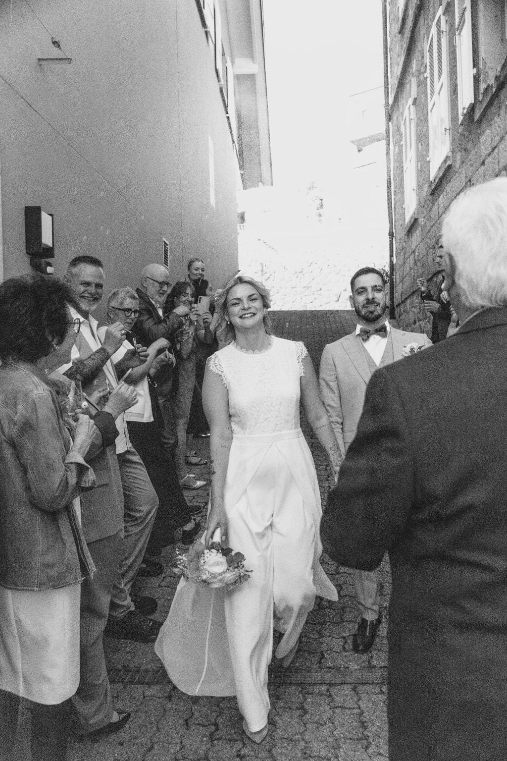 Kenza et Steven sortent sous les applaudissements de leurs invités dans une ruelle pavée, en noir et blanc